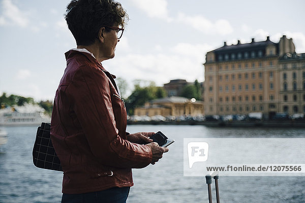 Seitenansicht einer älteren Frau  die ein Mobiltelefon hält  während sie in der Stadt am Meer steht.
