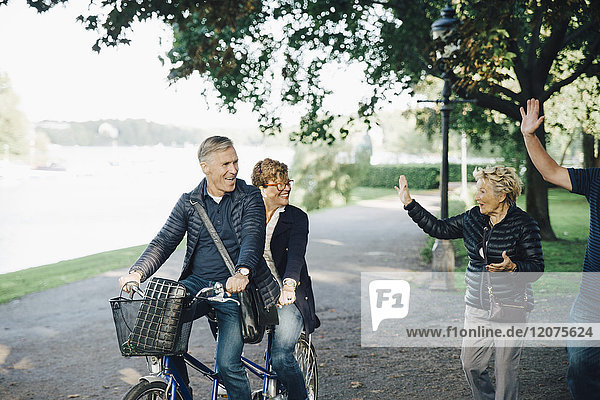 Senior couple waving friends riding tandem bike in park
