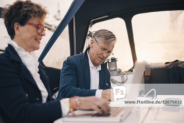 Senior Mann schreibt auf Papier  während er von einer Frau mit einem Laptop in der Yacht sitzt.