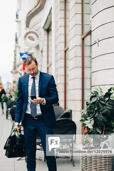 Reife Geschäftsmann mit Smartphone beim Gehen mit Tasche auf dem Bürgersteig in der Stadt