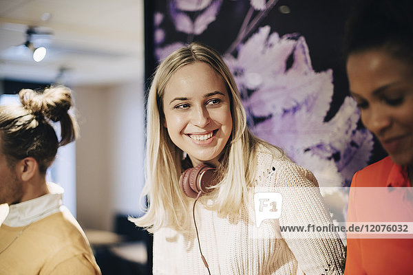 Lächelnde blonde Geschäftsfrau mit Kopfhörer von Geschäftsmann  die eine Kollegin im Büro anschaut.