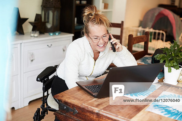 Lächelnde behinderte Frau spricht auf dem Handy  während sie sich den Laptop im Haus ansieht.