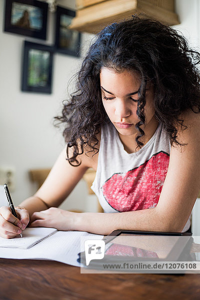 Teenager-Mädchen studiert am Tisch beim Schreiben in Buch durch digitale Tablette