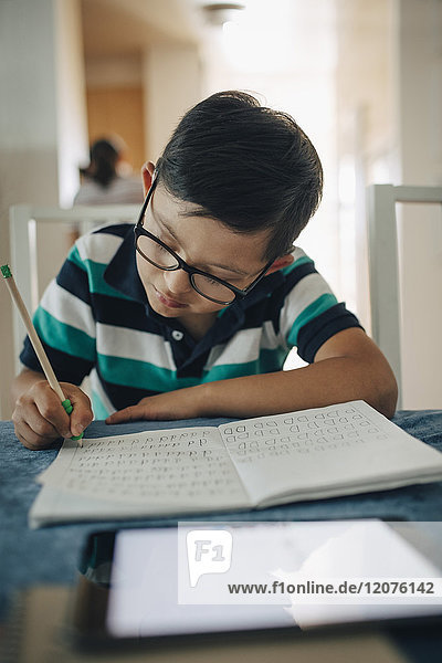 Nahaufnahme eines behinderten Jungen beim Schreiben auf einem Buch mit digitalem Tablett im Vordergrund am Tisch