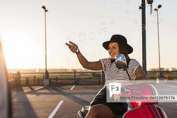 Lächelnde junge Frau auf dem Motorroller beim Spielen mit Seifenblasen