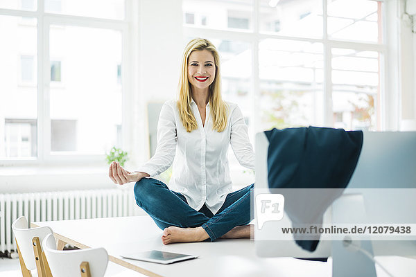 Lächelnde Geschäftsfrau sitzt auf dem Schreibtisch im Büro und übt Yoga.