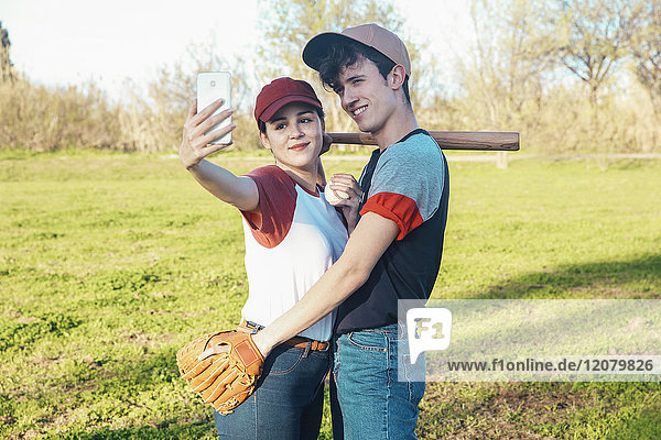 Lächelndes junges Paar mit Baseballausrüstung  das einen Selfie im Park nimmt.