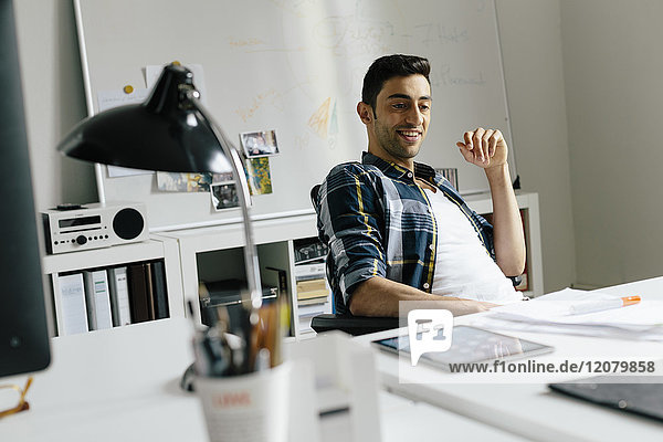 Lächelnder junger Mann mit Tablette und Notizen am Schreibtisch im Büro