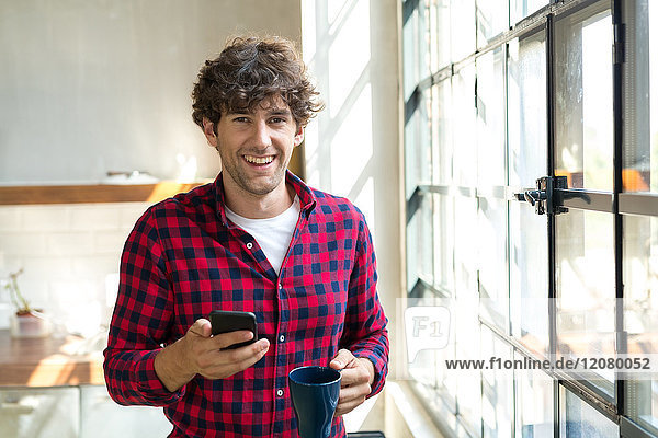 Jungunternehmer steht in der Firmenküche  trinkt Kaffee  nutzt Smartphone
