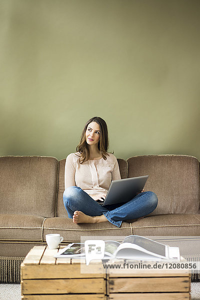 Lächelnde junge Frau sitzt auf der Couch mit Laptop