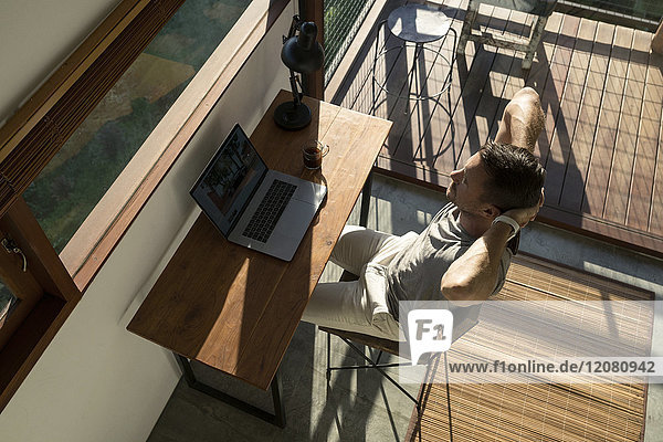 Man leaning back and taking a break from working at laptop on desk in house