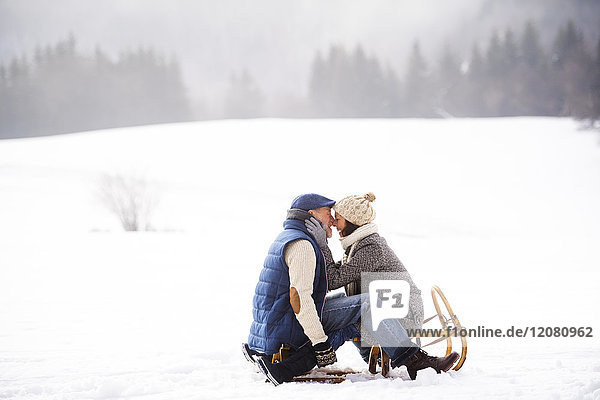 Happy senior couple sitting face to face on sledge in winter landscape