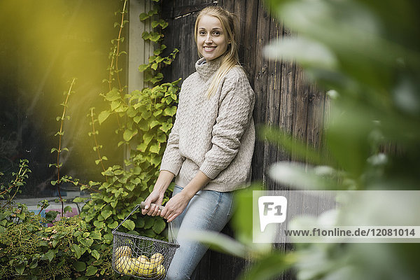 Portrait of smiling young woman holding wire basket with fruits in garden