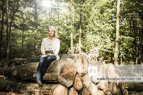 Porträt einer Frau  die auf einem Holzstapel im Wald mit Tablette sitzt.