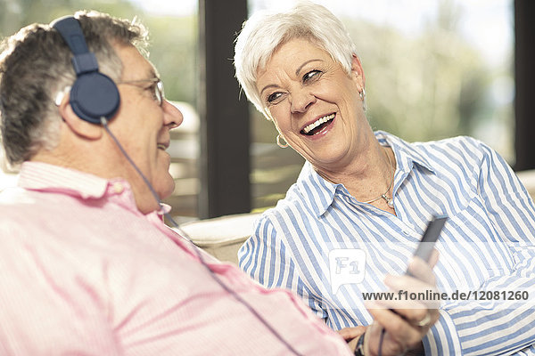 Glückliches Seniorenpaar mit Handy und Kopfhörer auf der Couch zu Hause