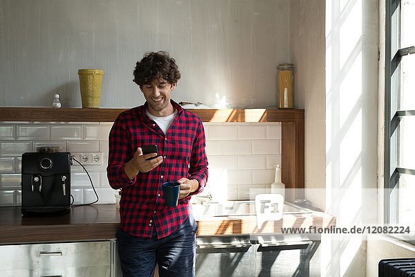 Young entrepreneur standing in company kitchen  drinking coffee  using smartphone