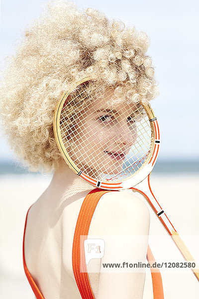 Porträt einer jungen Frau,  die durch das Fadenkreuz eines Tennisschlägers am Strand schaut.