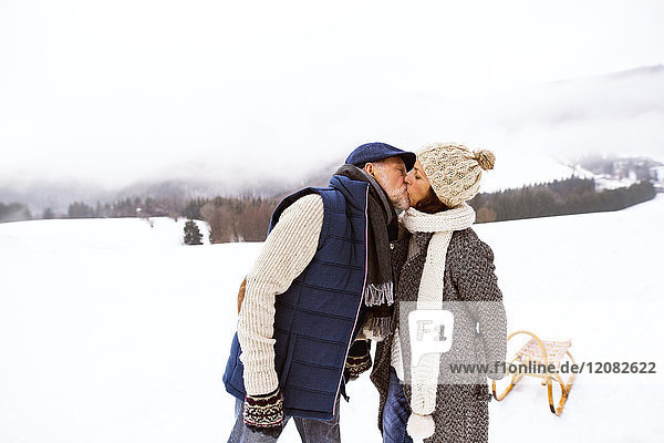 Küssendes Seniorenpaar mit Schlitten in verschneiter Landschaft