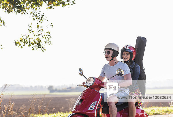Fröhliches junges Paar auf Motorroller mit Gitarrentasche auf dem Rücken