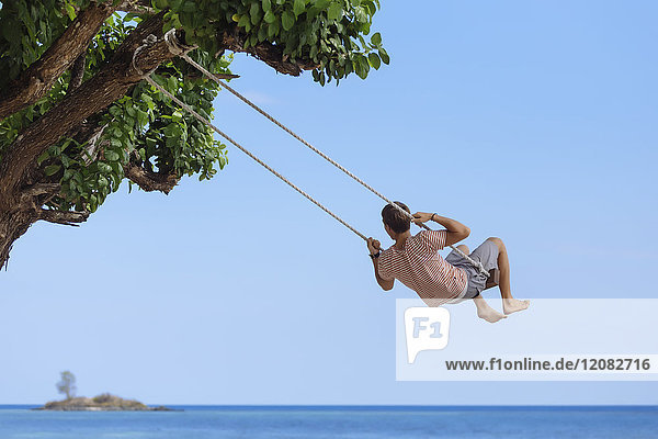 Indonesien, Insel Lombok, Mann auf einer Schaukel sitzend mit Blick auf die Ferne