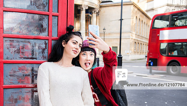 Two young stylish women taking smartphone selfie by red phone box  London  UK