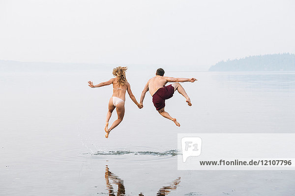 Young couple on beach  holding hands  jumping  mid air  rear view