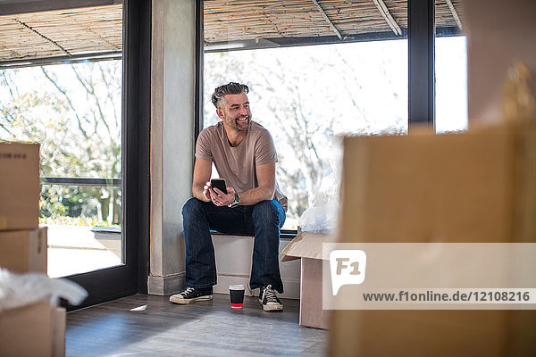 Man sitting in unfurnished home surrounded by cardboard boxes  using smartphone