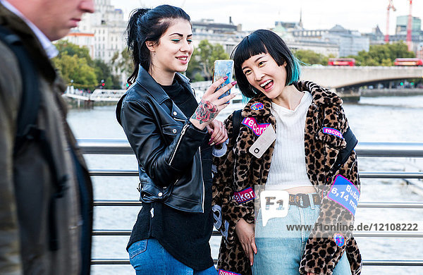 Zwei junge stilvolle Frauen lachen über Smartphones auf der Millennium-Fußgängerbrücke  London  Großbritannien