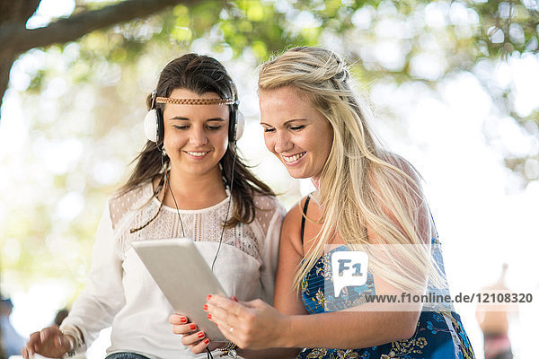 Zwei junge Freundinnen sitzen unter einem Baum  benutzen ein digitales Tablett und hören Kopfhörer