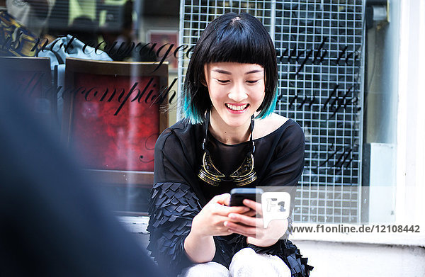 Junge elegante Frau sitzt vor einem Geschäft und schaut auf ein Smartphone