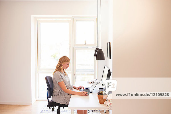 Schwangere junge Frau am Schreibtisch beim Tippen am Laptop