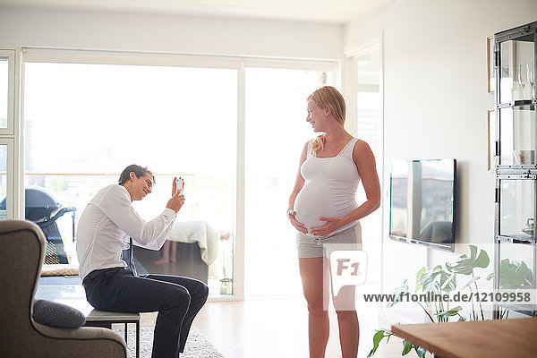 Man taking smartphone photograph of pregnant girlfriend in living room