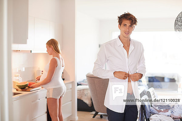 Portrait of man buttoning shirt and pregnant girlfriend in kitchen