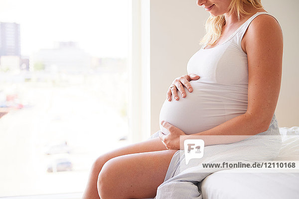 Schnappschuss einer schwangeren jungen Frau  die mit den Händen auf dem Bauch auf dem Bett sitzt