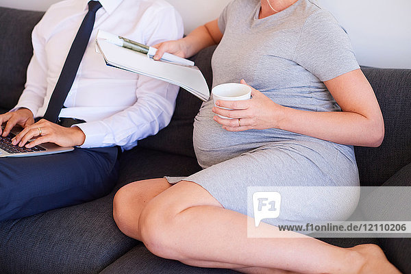 Nackenansicht eines schwangeren Paares  das mit Zeitschrift und Laptop auf dem Sofa sitzt