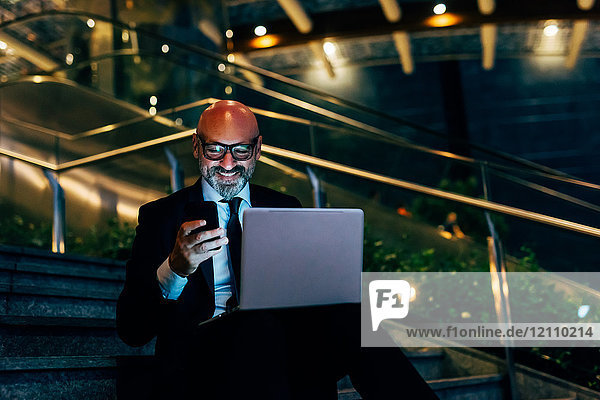 Reifer Geschäftsmann nachts im Freien  sitzt auf Treppen  benutzt Laptop  hält Smartphone