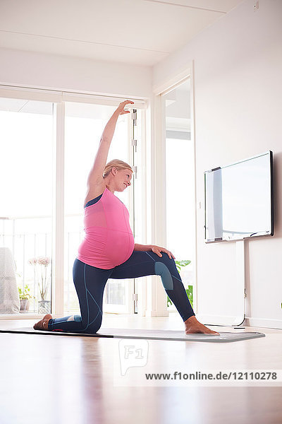 Schwangere junge Frau kniend bei einer Yoga-Übung im Wohnzimmer