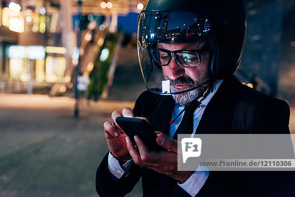 Mature businessman outdoors at night  wearing motorcycle helmet  using smartphone
