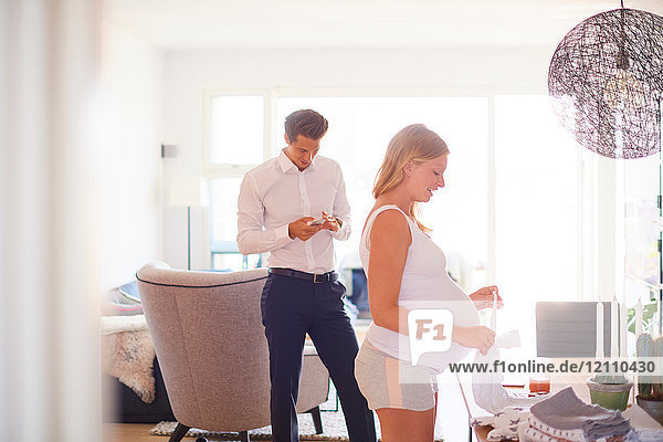 Man looking at smartphone and pregnant girlfriend folding laundry in living room