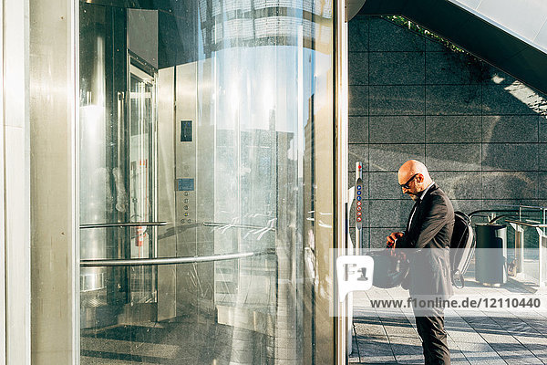Reifer Geschäftsmann im Freien  wartet auf den Aufzug  kontrolliert die Armbanduhr  hält den Motorradhelm