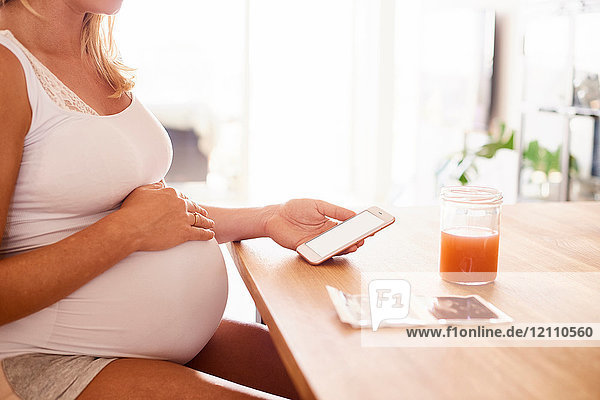 Ausschnittaufnahme einer schwangeren jungen Frau am Schreibtisch beim Betrachten von Smartphone- und Ultraschallbildern