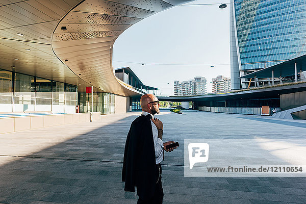 Reifer Geschäftsmann geht im Freien  trägt Anzugjacke über die Schulter  hält Smartphone