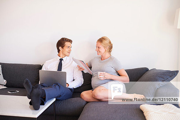 Auf dem Sofa sitzendes schwangeres Paar mit Zeitschrift und Laptop