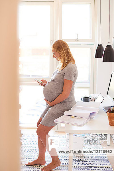 Schwangere junge Frau lehnt am Schreibtisch und schaut auf Smartphone