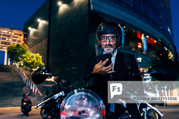 Reifer Geschäftsmann nachts im Freien  sitzt auf einem Motorrad und benutzt ein Smartphone