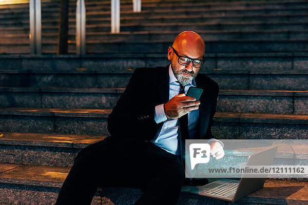 Älterer Geschäftsmann nachts im Freien  auf der Treppe sitzend  mit Smartphone  Laptop auf der Stufe neben ihm