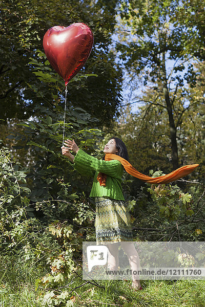 Lächelnde Frau hält roten Herzballon und steht auf dem Feld gegen Bäume im Park