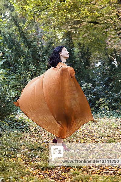 Volle Länge der Frau mit orangem Schal  die im Park gegen Bäume tanzt.