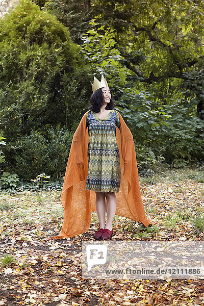 Volle Länge der lächelnden Frau mit Krone  die im Herbst gegen Bäume im Park steht.