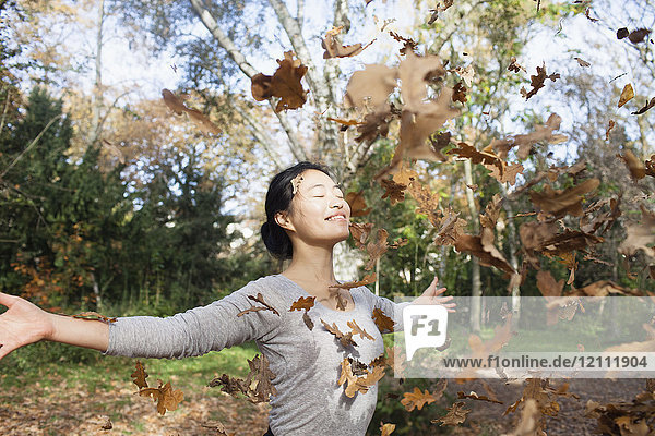 Trockene Blätter fallen auf eine glückliche Frau  die im Herbst mit ausgestreckten Armen im Park steht.
